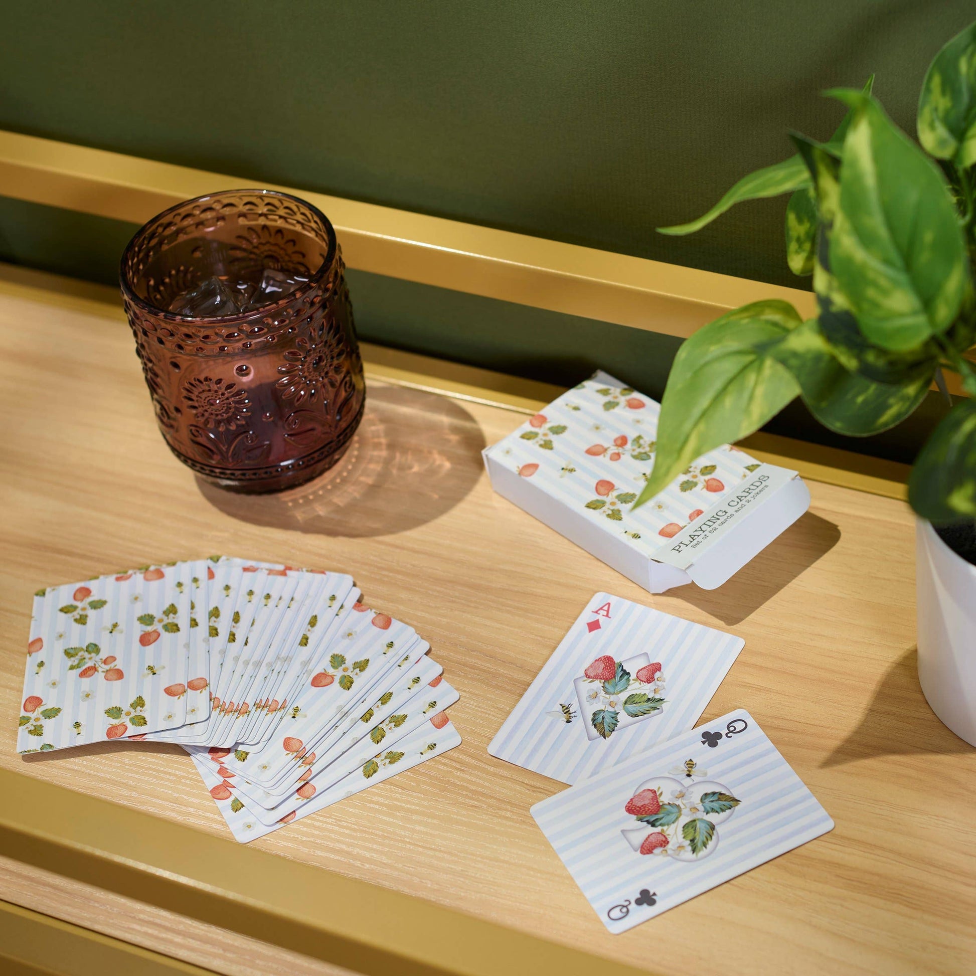 Playing cards with floral design on a wooden surface with a glass and plant in the background.